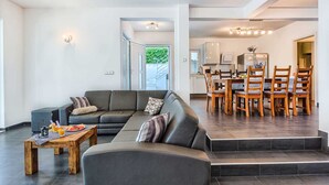 Un salon moderne avec un canapé d’angle, une table basse en bois sur laquelle sont disposés des fruits et des boissons, et un coin repas avec des chaises et une table en bois.