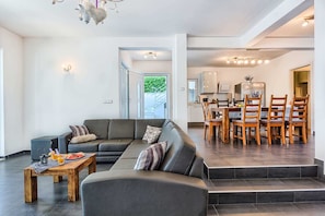 Un salon moderne avec un canapé d’angle, une table basse en bois sur laquelle sont disposés des fruits et des boissons, et un coin repas avec des chaises et une table en bois.
