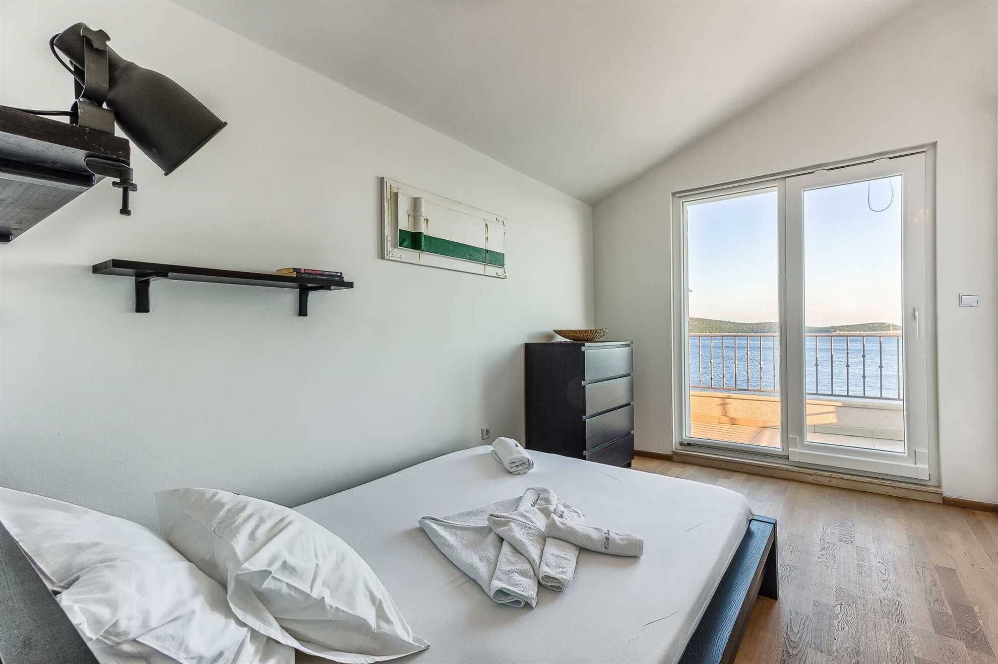 A bedroom with a bed, a desk, a chair, a shelf, a picture, and a view of the sea through a sliding door.