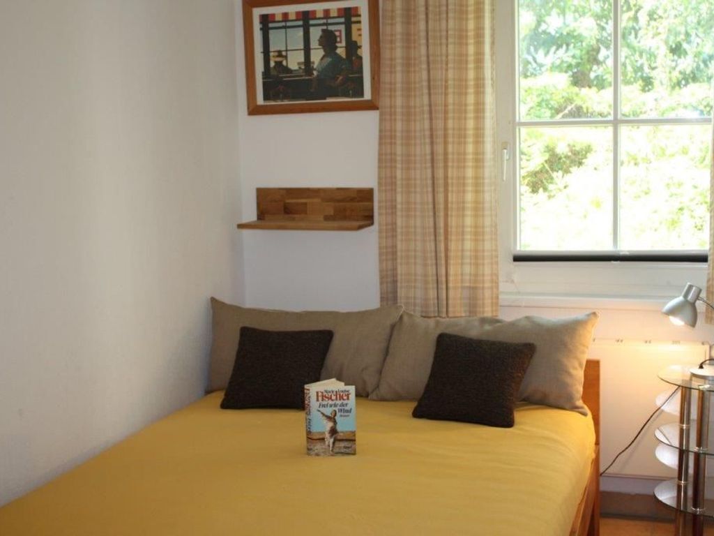 A bed with a yellow bedspread, two pillows, and a book titled "Frei da Gora". There is a window with curtains, a wall-mounted shelf, and a lamp on a side table.