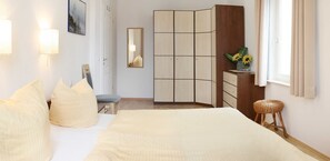 A bedroom with a bed, pillows, a bedside table, a mirror, a wardrobe, and a window with curtains.