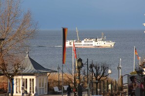 A coastal scene with a ferry named 'ADLER-VINETA' in the water, a small building with a clock, and flags.