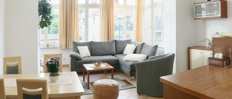 A modern living room with a grey sectional sofa, a wooden dining table, and a kitchen area with a wooden counter and cabinets.