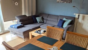 A modern living room with a sofa, dining table, wicker chairs, and artwork on the wall. - Vacation home Boddenblick (Barth)