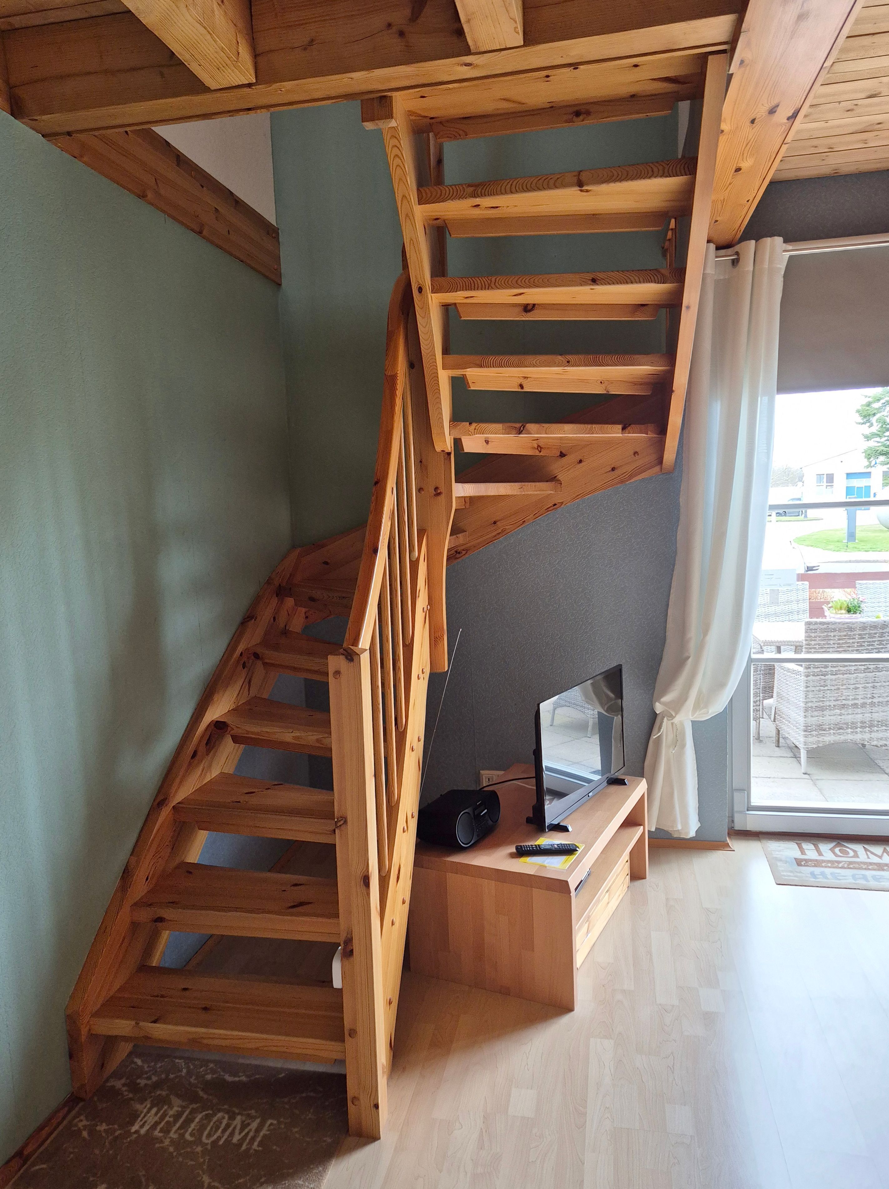 Eine Holztreppe führt zu einer erhöhten Wohnfläche mit einem Flachbildfernseher und einem Lautsprecher auf einer Holzkonsole.