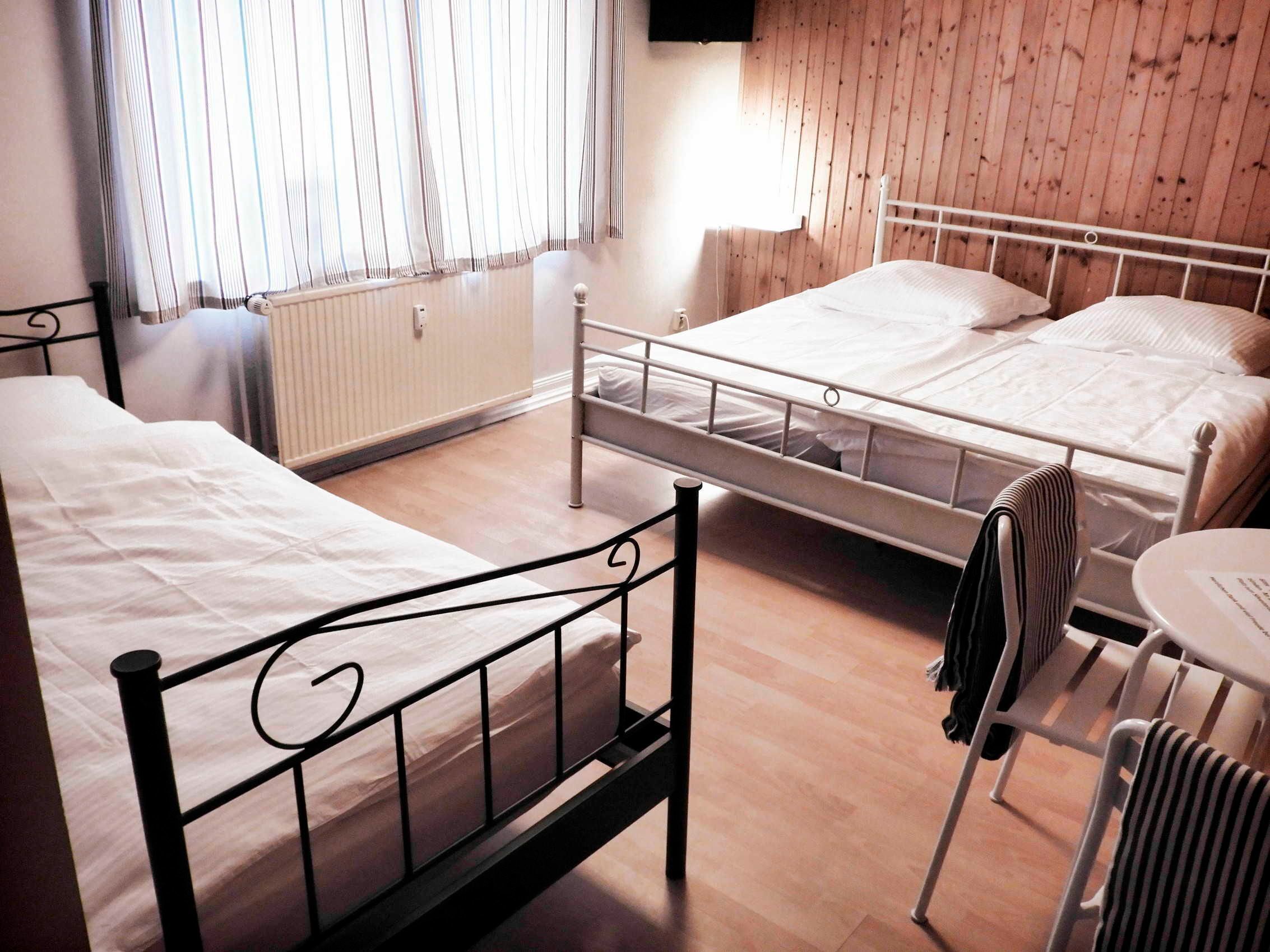 Quarto com duas camas, um radiador, janela com cortinas e uma pequena mesa com cadeira.