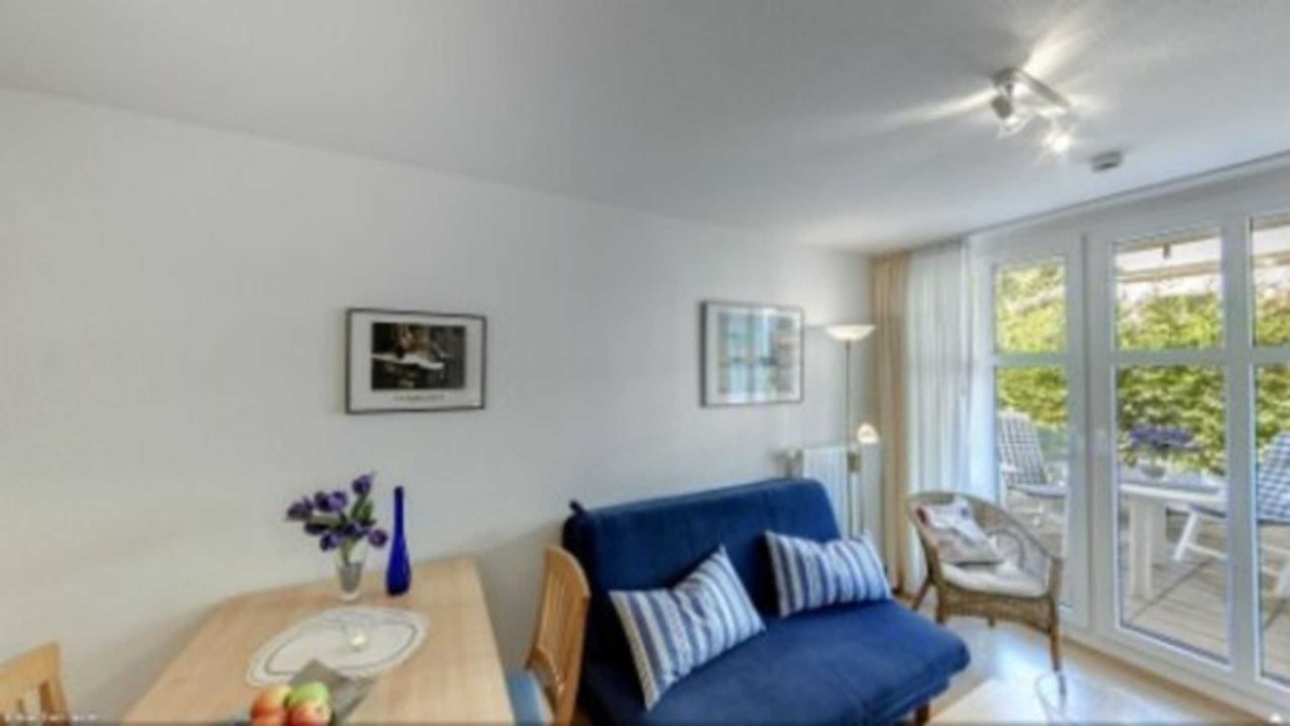 A modern living room with a blue sofa, a dining table with a vase of flowers, and a large window with a view of greenery.