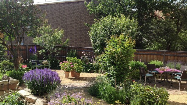 A garden with a patio area, stone wall, and various plants.