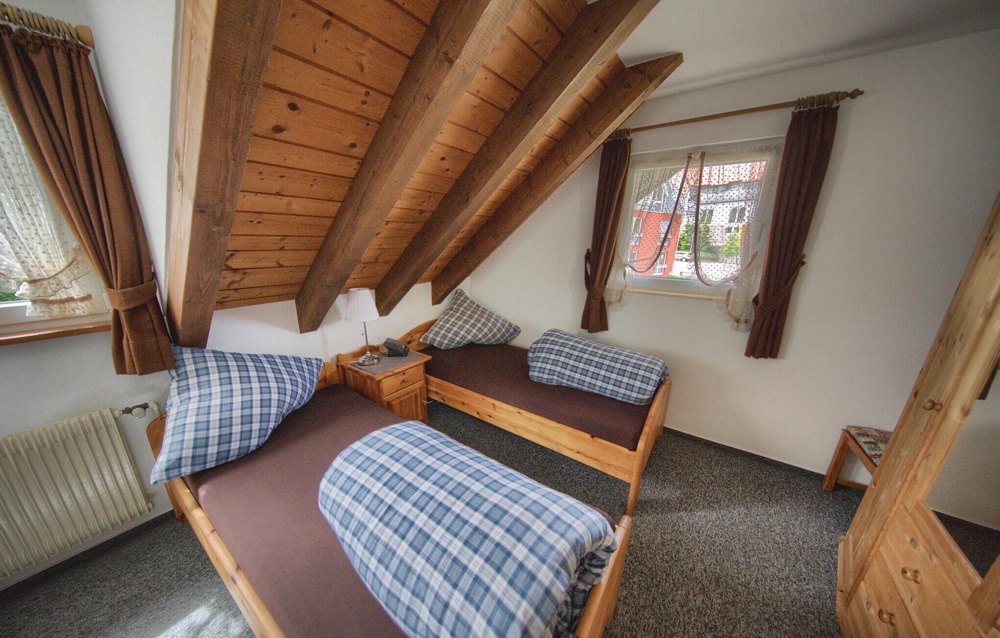 Una habitación pequeña y acogedora con techo inclinado, una cama individual con colcha de cuadros, una mesita de noche con lámpara y una ventana con cortinas.