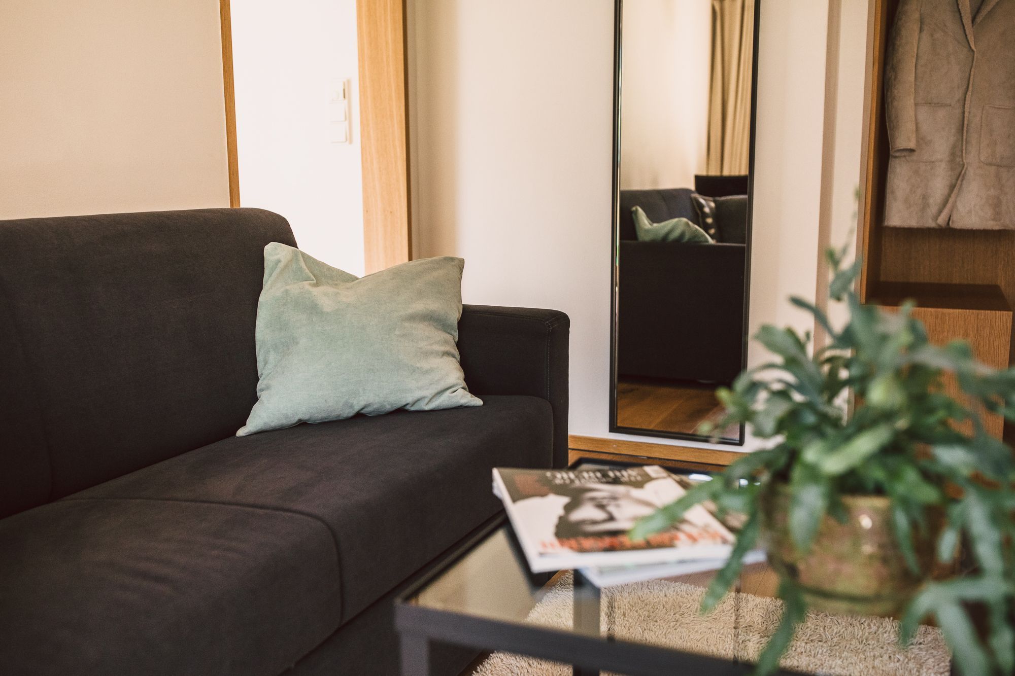 Ein Wohnzimmer mit einem dunklen Sofa, einem hellgrünen Kissen, einer Zeitschrift auf einem kleinen Tisch und einem großen Spiegel, der den Raum reflektiert.