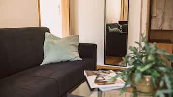 Ein Wohnzimmer mit einem dunklen Sofa, einem hellgrünen Kissen, einer Zeitschrift auf einem kleinen Tisch und einem großen Spiegel, der den Raum reflektiert.