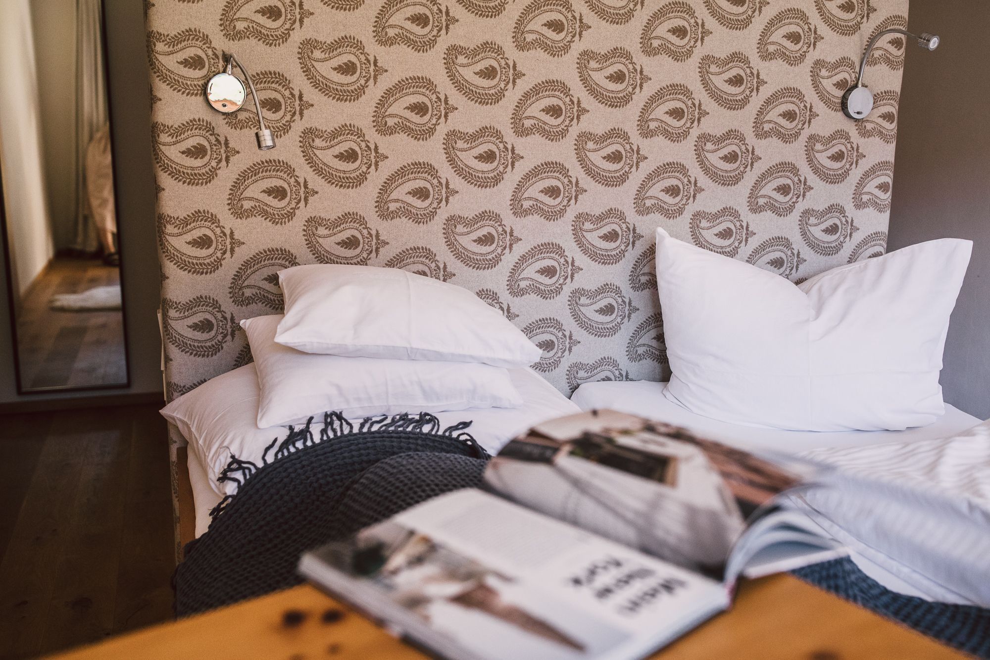 A bed with white pillows and a patterned headboard, a wooden nightstand with a magazine, and a mirror on the wall.