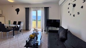 Ein modernes Wohnzimmer mit Sofa, Fernseher, Essbereich mit Stühlen und einem Balkon mit Vorhängen.