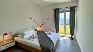 Ein Hotelzimmer mit Bett, Nachttisch mit Lampe, Fenster mit Vorhängen und Blick aufs Meer.
