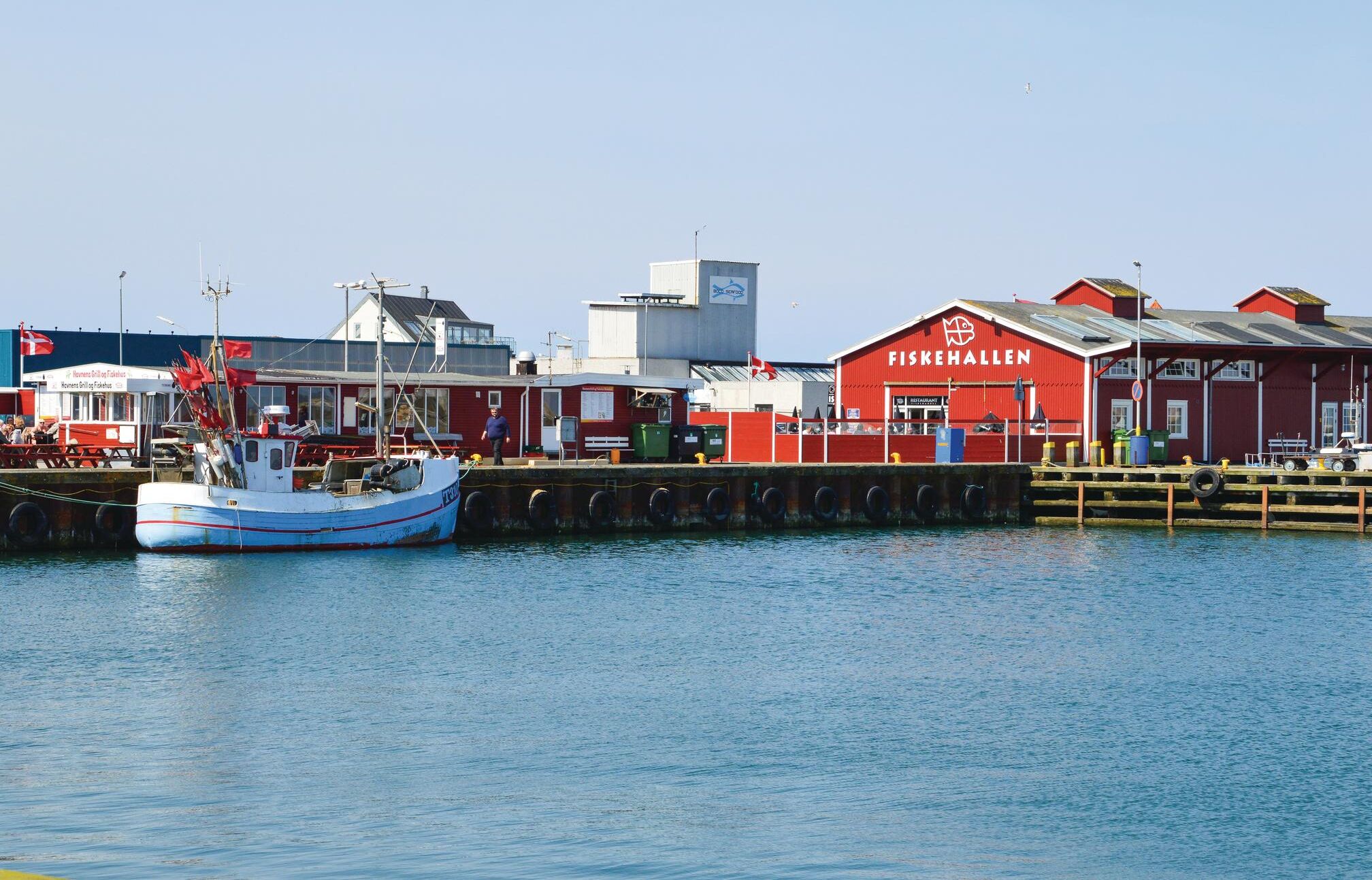 Un port avec un bâtiment rouge portant l’inscription « FISKEHALLEN » et un bateau amarré.
