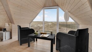 Habitación espaciosa con techos altos, una ventana grande, muebles de mimbre y una mesa con un periódico y un vaso.