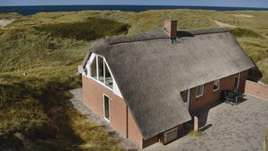 Una casa con techo de paja, con un ventanal grande y una puerta, ubicada sobre una duna cubierta de hierba y vistas al mar.