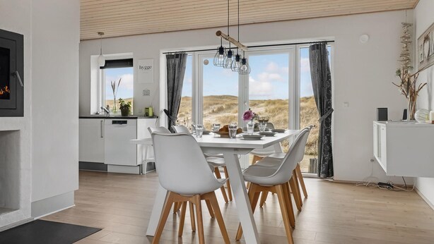 Una cocina moderna con muebles blancos, un comedor con sillas blancas y una mesa de madera, y ventanas amplias con vistas a las dunas.