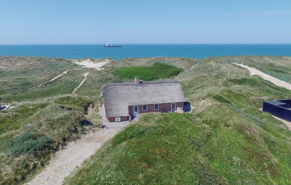 Una casa con techo de paja, con un camino de grava que la lleva a ella, rodeada de dunas cubiertas de hierba y vista al mar.