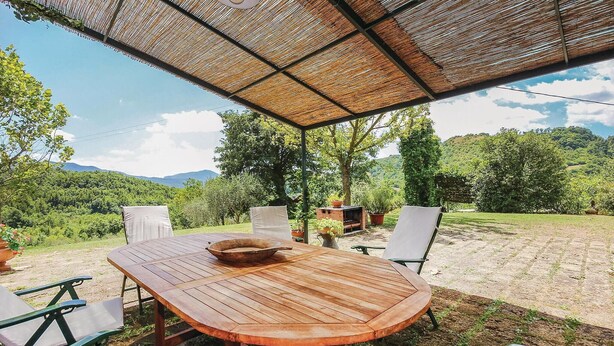 Una mesa de madera al aire libre con sillas, un toldo tejido en la parte superior y una vista panorámica de un paisaje exuberante con árboles y colinas al fondo.