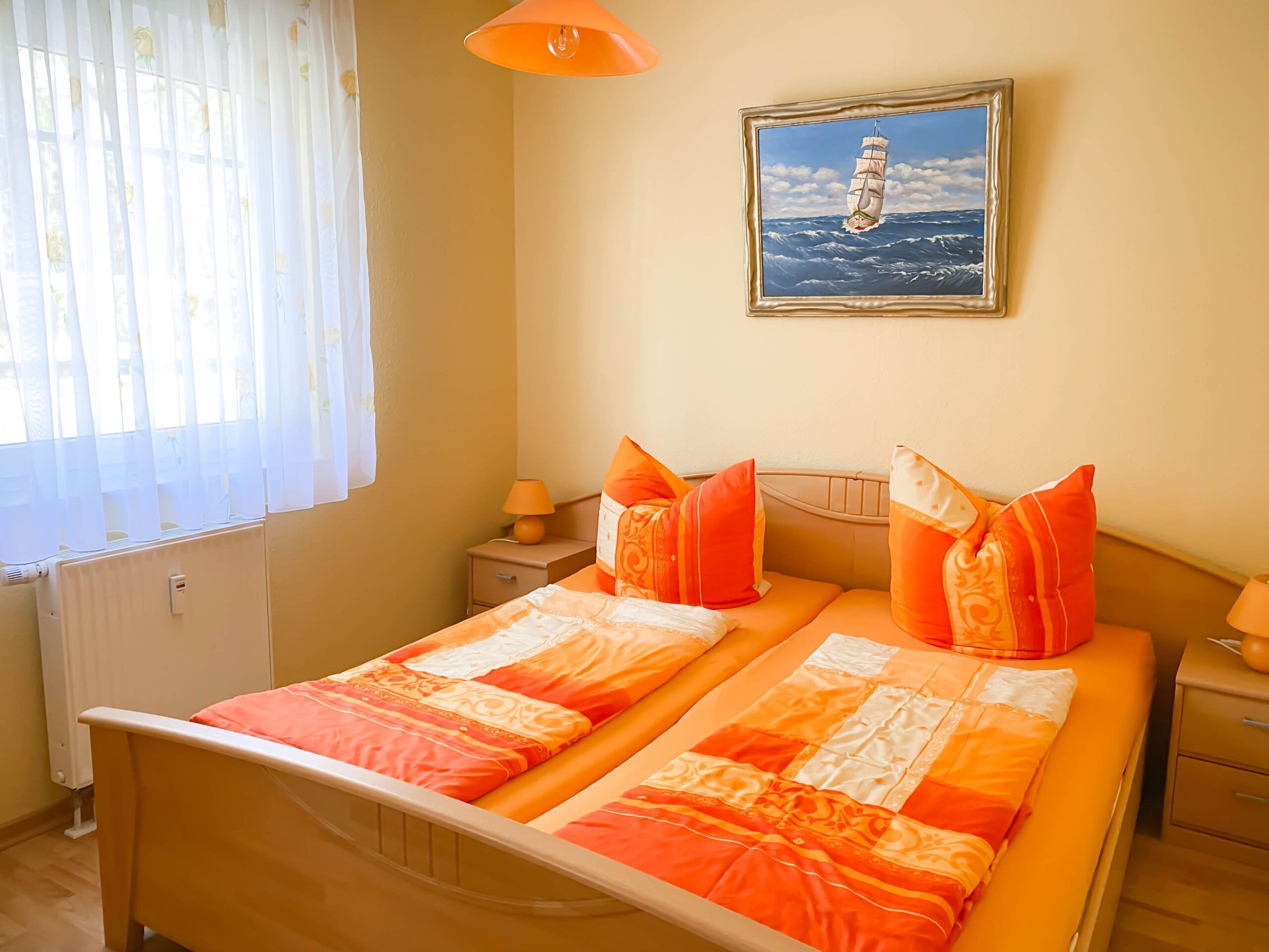 Cama doble con ropa de cama naranja, mesita de noche con lámpara, cuadro enmarcado de un velero en la pared y ventana con cortinas traslúcidas.