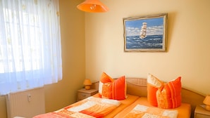 A double bed with orange bedding, a nightstand with a lamp, a framed picture of a sailboat on the wall, and a window with sheer curtains. - *Forest 10 measure GM 69552 - Forest 10 - SNR: 1 (Graal-Müritz)