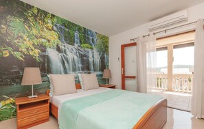 Une chambre avec un grand décor mural représentant une cascade, un lit recouvert d’une couverture verte, des tables de chevet en bois, deux lampes et une porte-fenêtre coulissante donnant sur un balcon.