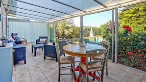 Une terrasse couverte avec une table ronde et des chaises, une maquette de voilier et une vue sur un jardin parsemé de fleurs rouges.