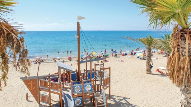 A beach with a wooden play structure, palm trees, and a clear blue sea. - Cozy apartment in Alicante with WiFi (Alicante)