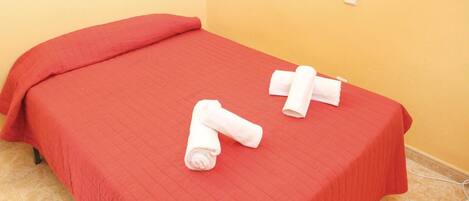 A small bed with a red bedspread and two rolled towels on it.