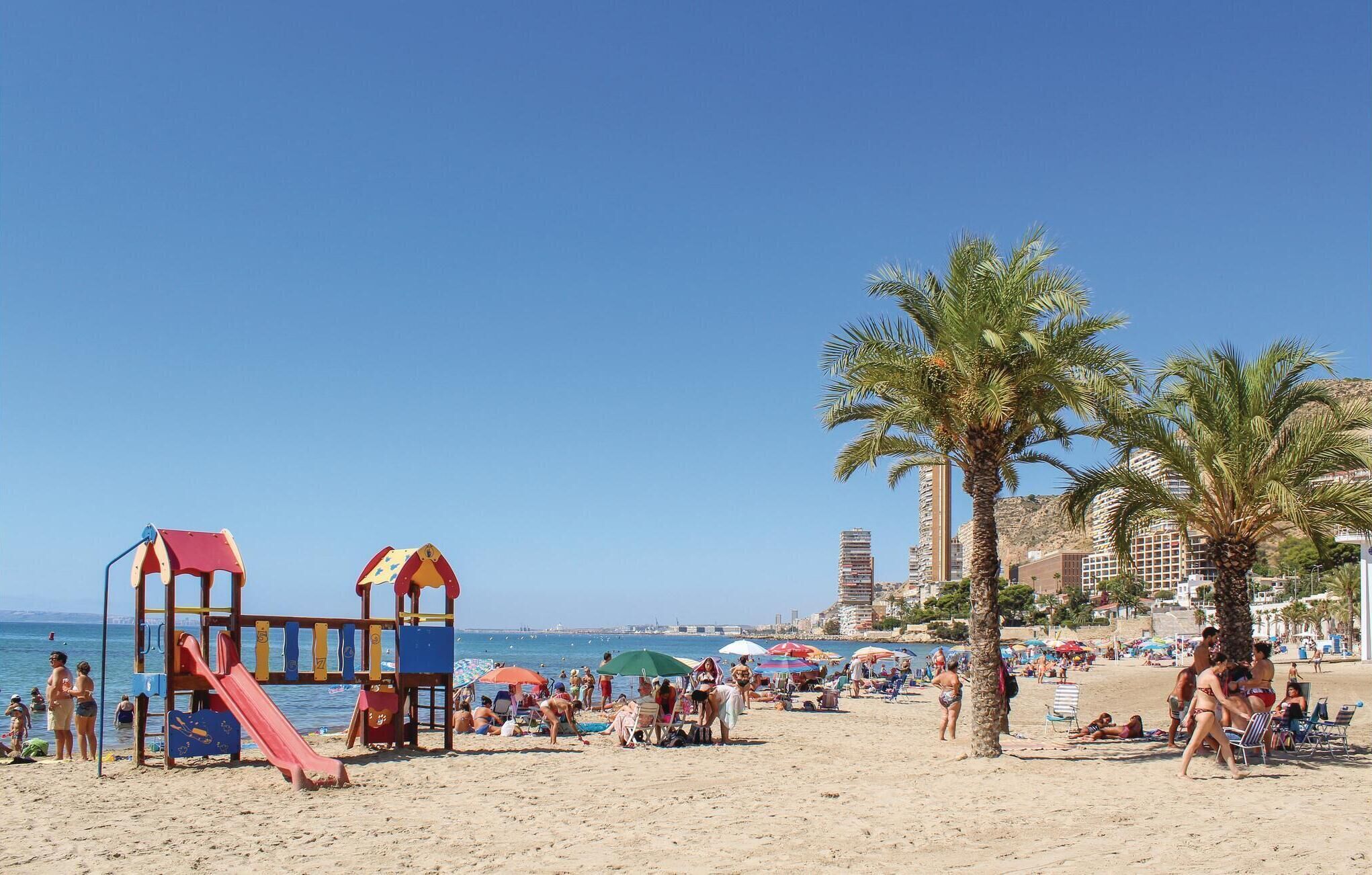 Une plage avec une aire de jeux, des palmiers et une vue sur la mer et les bâtiments en arrière-plan.