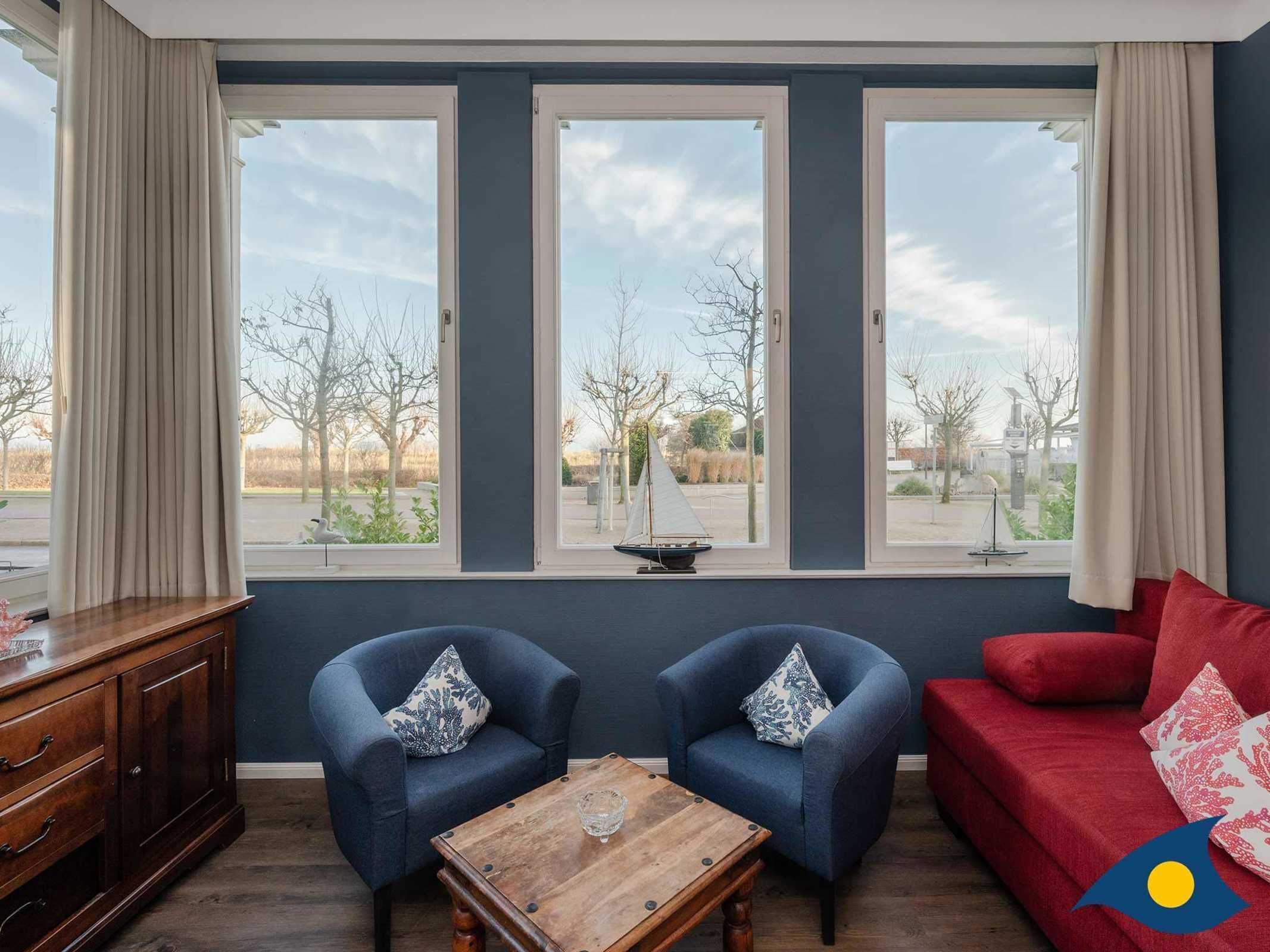 Een kamer met een groot raam, twee blauwe fauteuils, een houten bijzettafeltje en een rode bank.