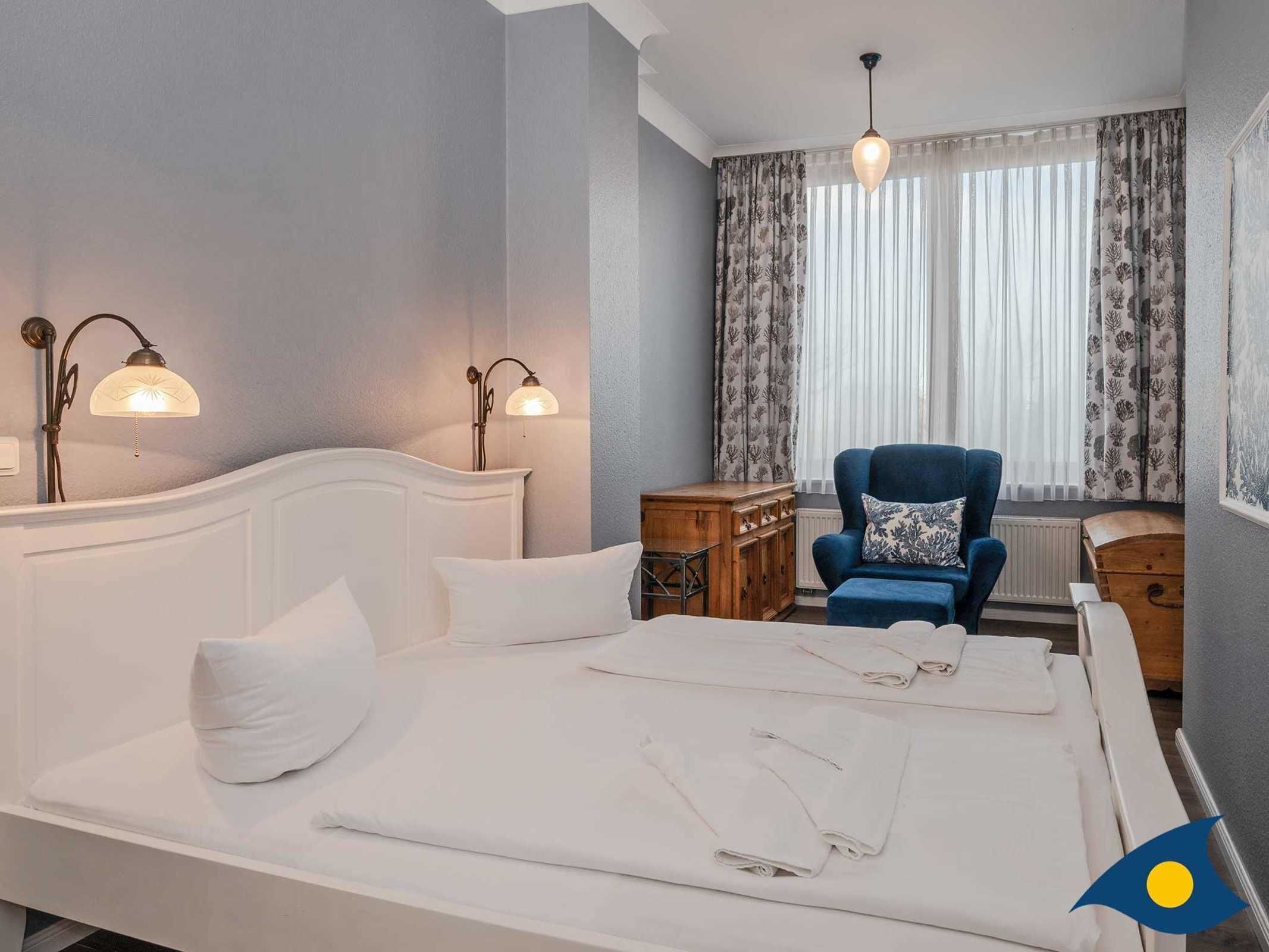 A hotel room with a bed, two wall-mounted lamps, a blue armchair, a wooden nightstand, and a window with sheer curtains.