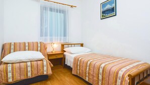A bedroom with two beds, a wooden headboard, a bedside table with a lamp, a window with sheer curtains, and a framed picture on the wall.