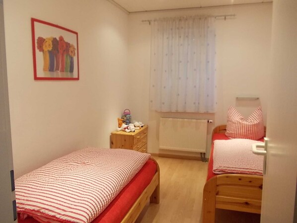 Un dormitorio pequeño y ordenado con dos camas individuales, un aparador de madera, un radiador y una ventana con cortinas traslúcidas.