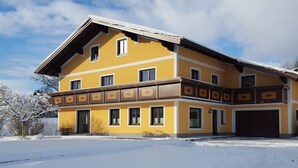A two-story building with a garage, a balcony with decorative trim, and a snow-covered ground. - Apartment Schafberg - Gästehaus Wienerroither (Mondsee)