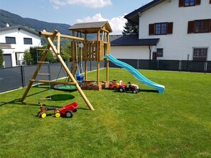 Un parque infantil con tobogán y estructuras para escalar, rodeado por una zona vallada y un césped.