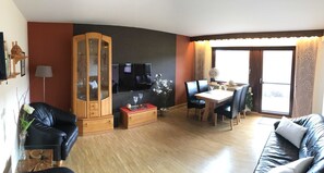 A living room with a black leather sofa, a wooden cabinet, a dining table with chairs, and a TV.
