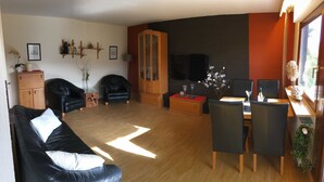 A living room with a black leather sofa, two black armchairs, a wooden cabinet, a TV, and a dining area with a table and chairs.