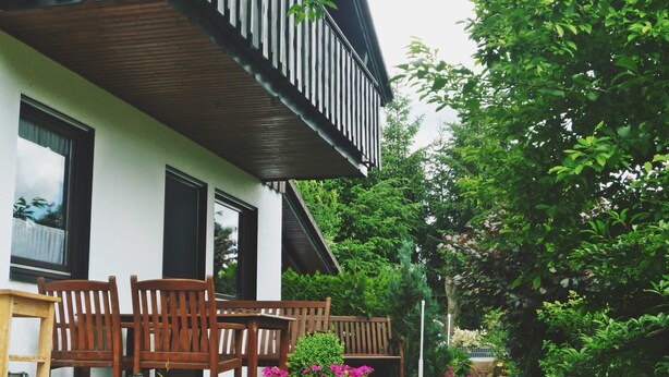 A house with a balcony, wooden chairs, and a garden with purple flowers.