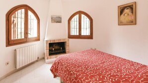 A bedroom with a fireplace, two arched windows, a bed with a red floral bedspread, and a painting on the wall. - Awesome home in Calonge (Calonge)