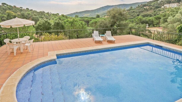 A swimming pool with a tiled edge, surrounded by a tiled deck and a metal railing. There are white lounge chairs and a table with chairs, and an umbrella. The background features a hilly landscape with trees and buildings. - Awesome home in Calonge (Calonge)