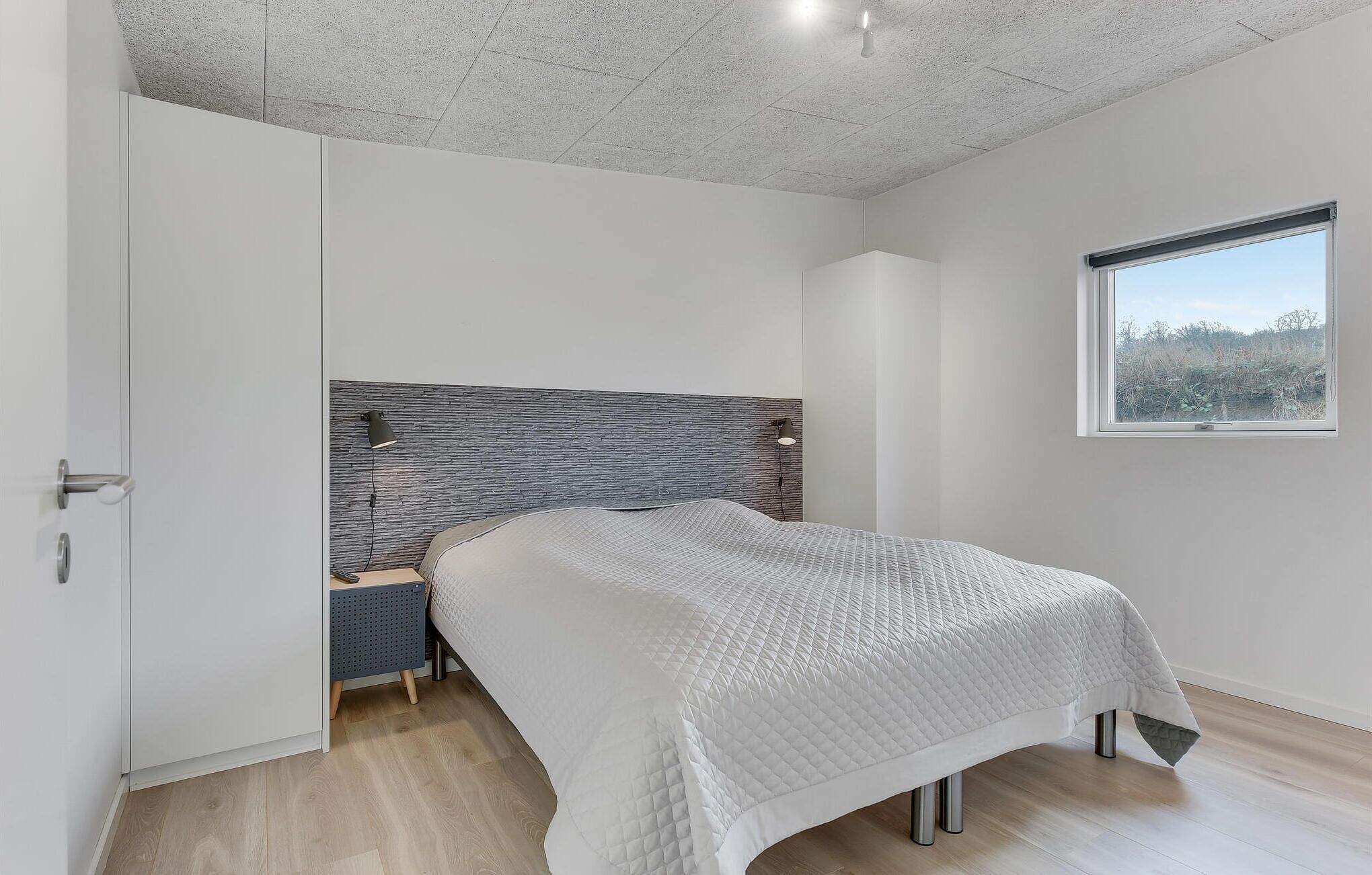 A bedroom with a bed, a nightstand, a wall-mounted lamp, and a window with a view of trees.