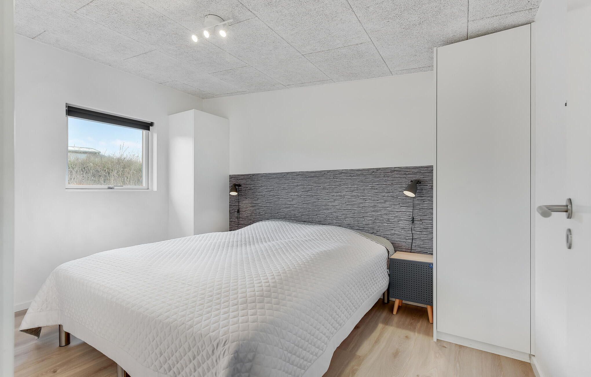 A modern bedroom with a large bed, a headboard with a textured design, a small bedside table, and a window with a view of the outdoors.