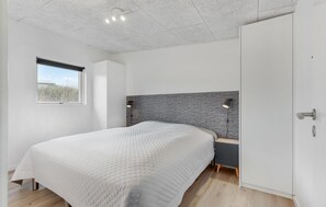 Une chambre à coucher moderne avec un grand lit, une tête de lit au motif texturé, une petite table de chevet et une fenêtre donnant sur l’extérieur.