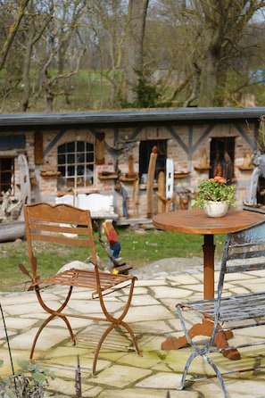 A rustic outdoor seating area with a wooden table and metal chairs, a potted plant, and a brick building in the background.