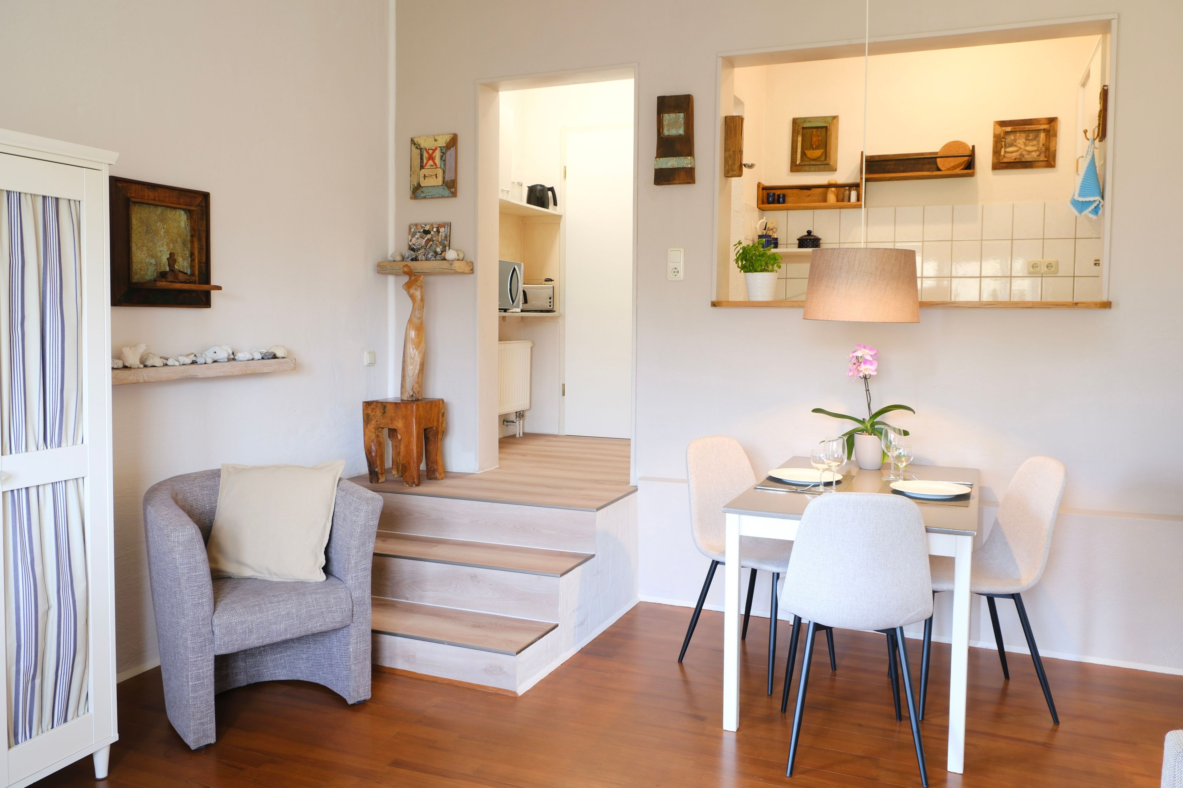 Ein modernes Wohnzimmer mit einem grauen Sessel, einem Esstisch mit Stühlen, einer Treppe und einem durchgehenden Türspalt sichtbaren Küchenbereich.