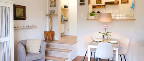 Una sala moderna con un sillón gris, una mesa de comedor con sillas, una escalera y una cocina visible a través de una puerta abierta.
