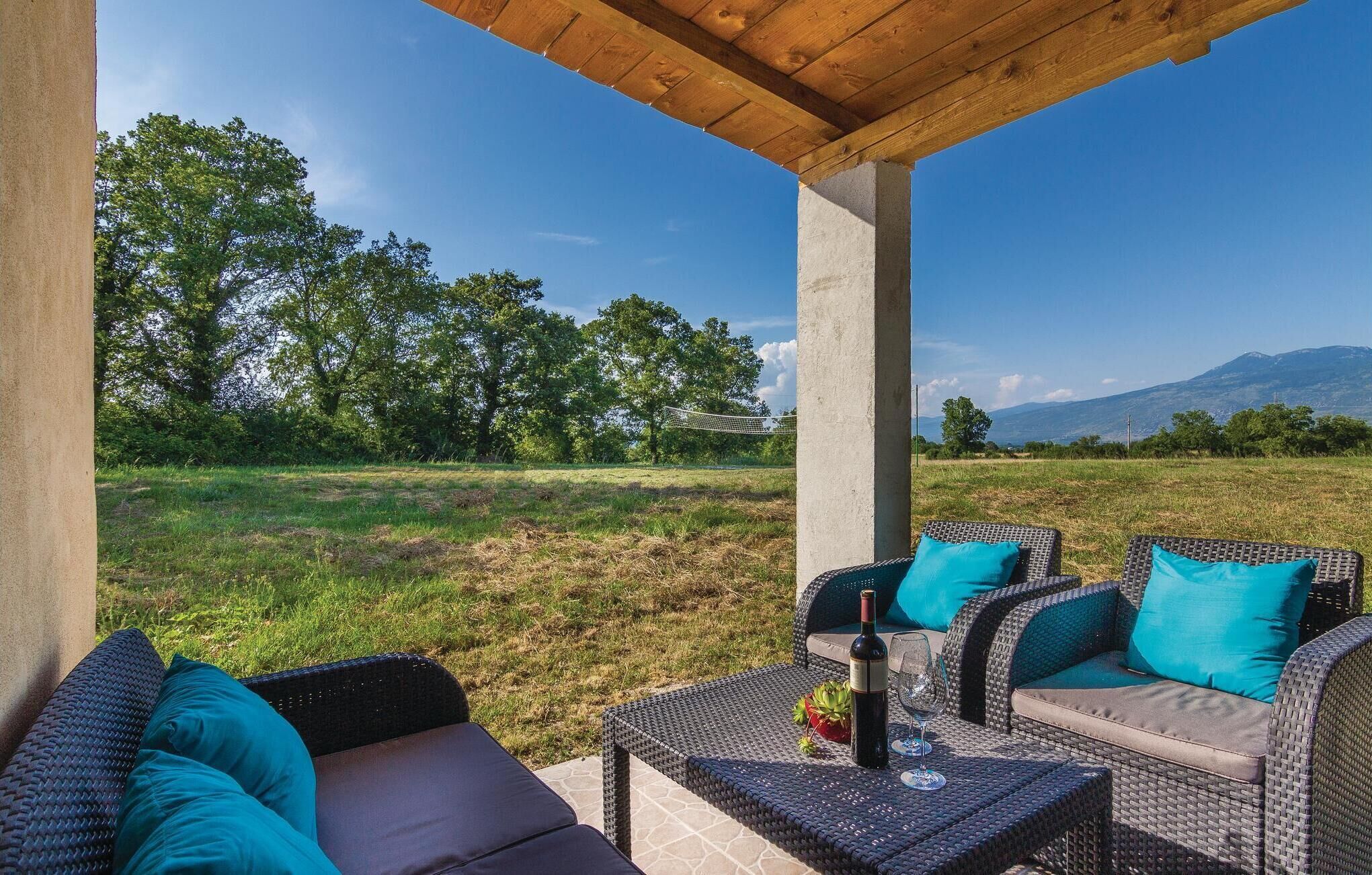 Ein Außenbereich mit Korbmöbeln und Kissen, ein Tisch mit einer Flasche und Gläsern sowie Blick auf ein grünes Feld und Berge im Hintergrund.