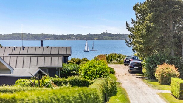 A coastal road with parked cars, a sailboat on the water, and a modern building with a sloped roof. - 3 bedroom amazing home in Egernsund (Egernsund)